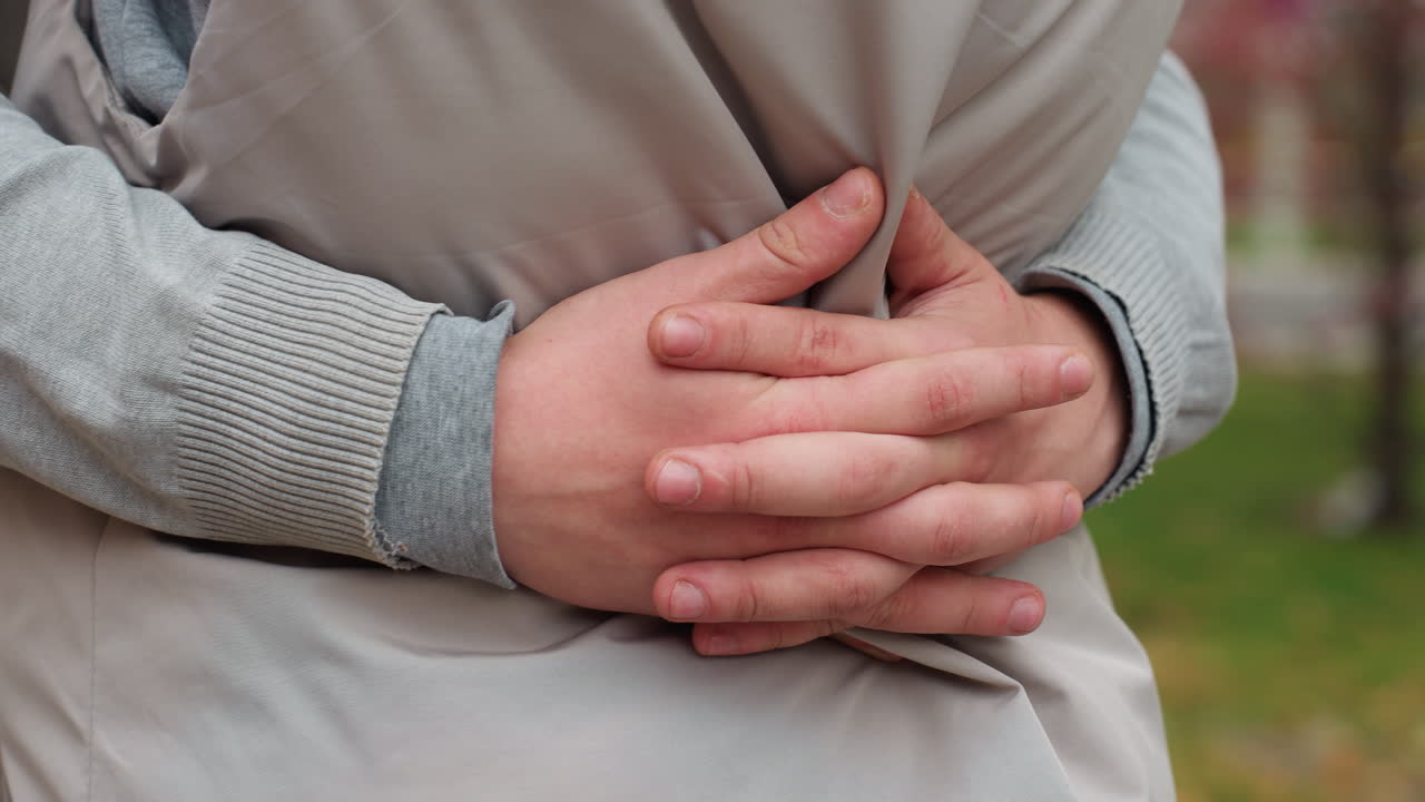 Close up of lovers embracing tightly around waist, hand resting gently on jacket, showing affection and intimacy, with soft background featuring blurred figure passing by on scooter