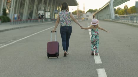 mother and daughter walking outdoors to airport. woman carrying suitcase bag. child and mom vacation