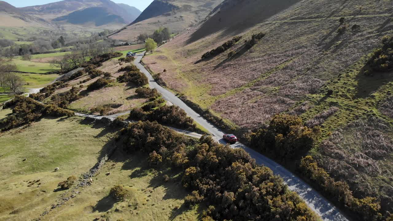 Aerial footage of a red Nissan Qashqai traveling on a winding road through the picturesque Lake District landscape. Features hilly terrain, lush greenery, and sunlight casting long shadows.