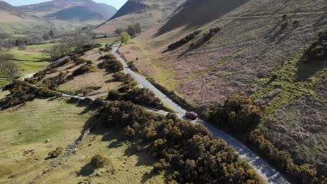 aerial footage of a red nissan qashqai traveling on a winding road through the picturesque lake district landscape. features hilly terrain, lush greenery, and sunlight casting long shadows.