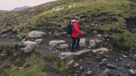 un joven con chaqueta roja y una mochila negra está cruzando un pequeño río en la ladera de la montaña en su caminata a cámara lenta en las islas feroe