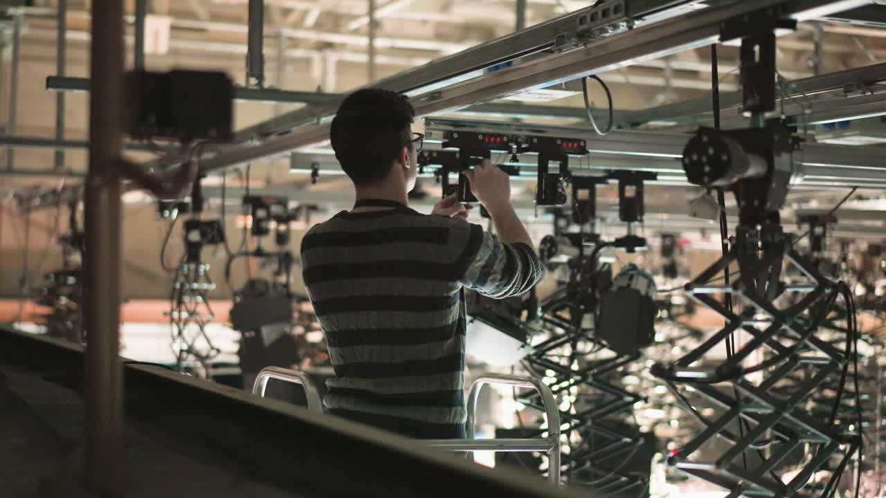 Worker adjusting stage lighting equipment on elevated platform in large studio. Focus on technician handling controls for overhead lights during production setup. High-angle shot of complex lighting grid