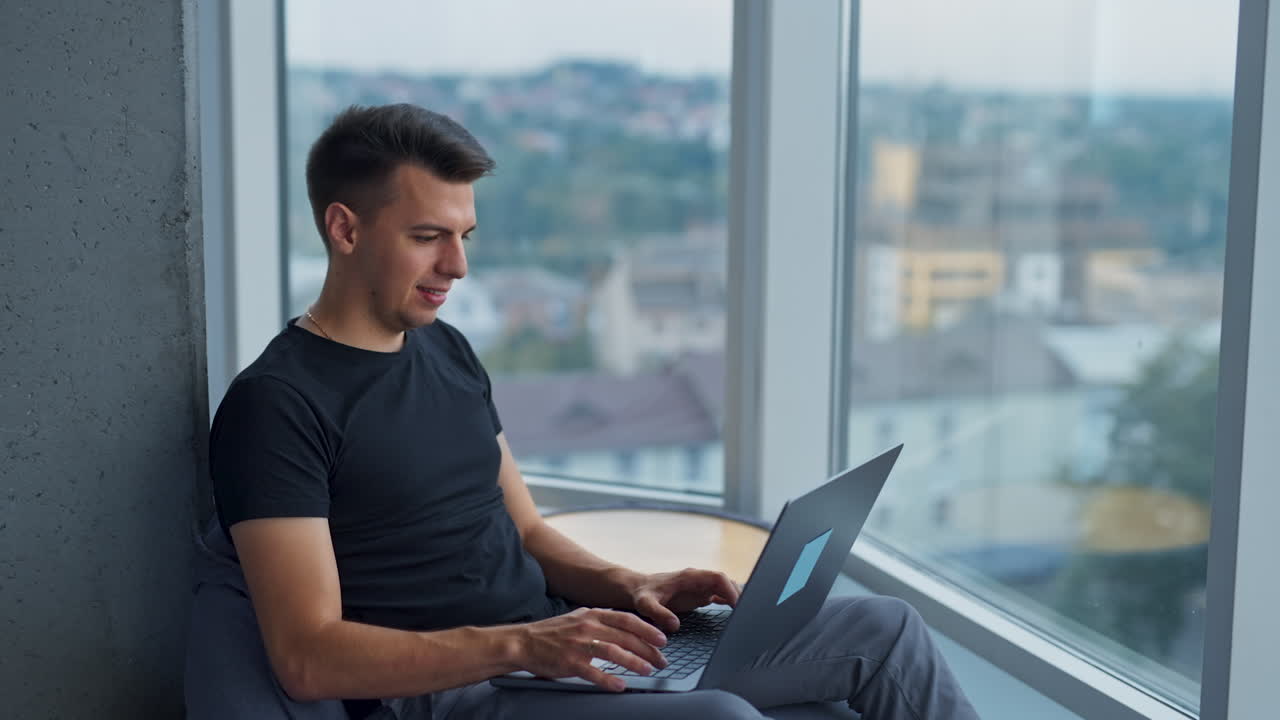 Young creative handsome freelancer working at computer. Male self-employed entrepreneur working sitting in bean bag chair near window. Blurred backdrop.