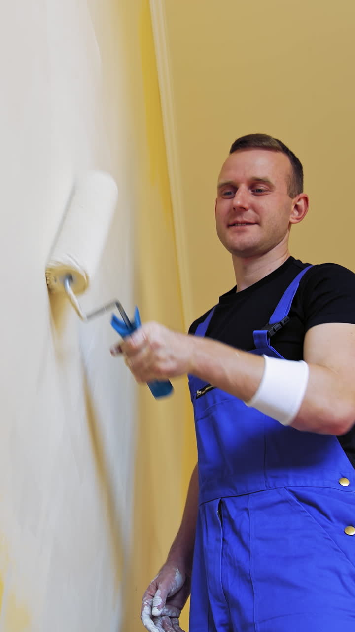 Roller repainting wall into light color. Young man in blue overalls painting wall into white in the apartment. Makeover in flat. Vertical video