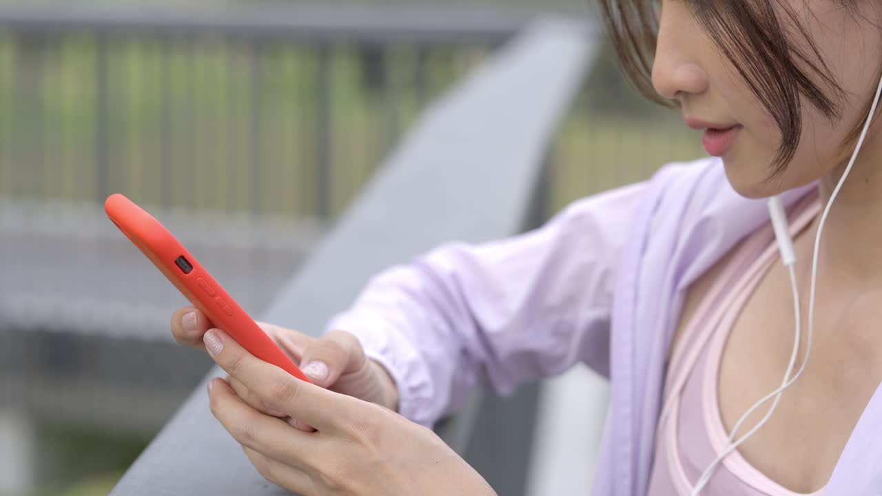 4k 젊은 아시아 여성이 아침에 공원에서 조하는 동안 이어폰과 스마트폰에서 음악을 듣고 있습니다.