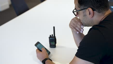 una persona, con gafas y una camisa negra, está usando un teléfono inteligente y un walkie-talkie colocado en una mesa blanca, posiblemente para tareas de comunicación o coordinación
