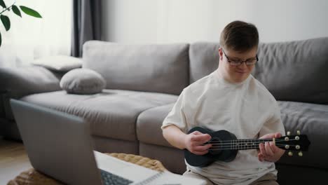 Down-syndrome-guy-having-fun-while-playing-ukulele,-learning-how-to-play-with-laptop