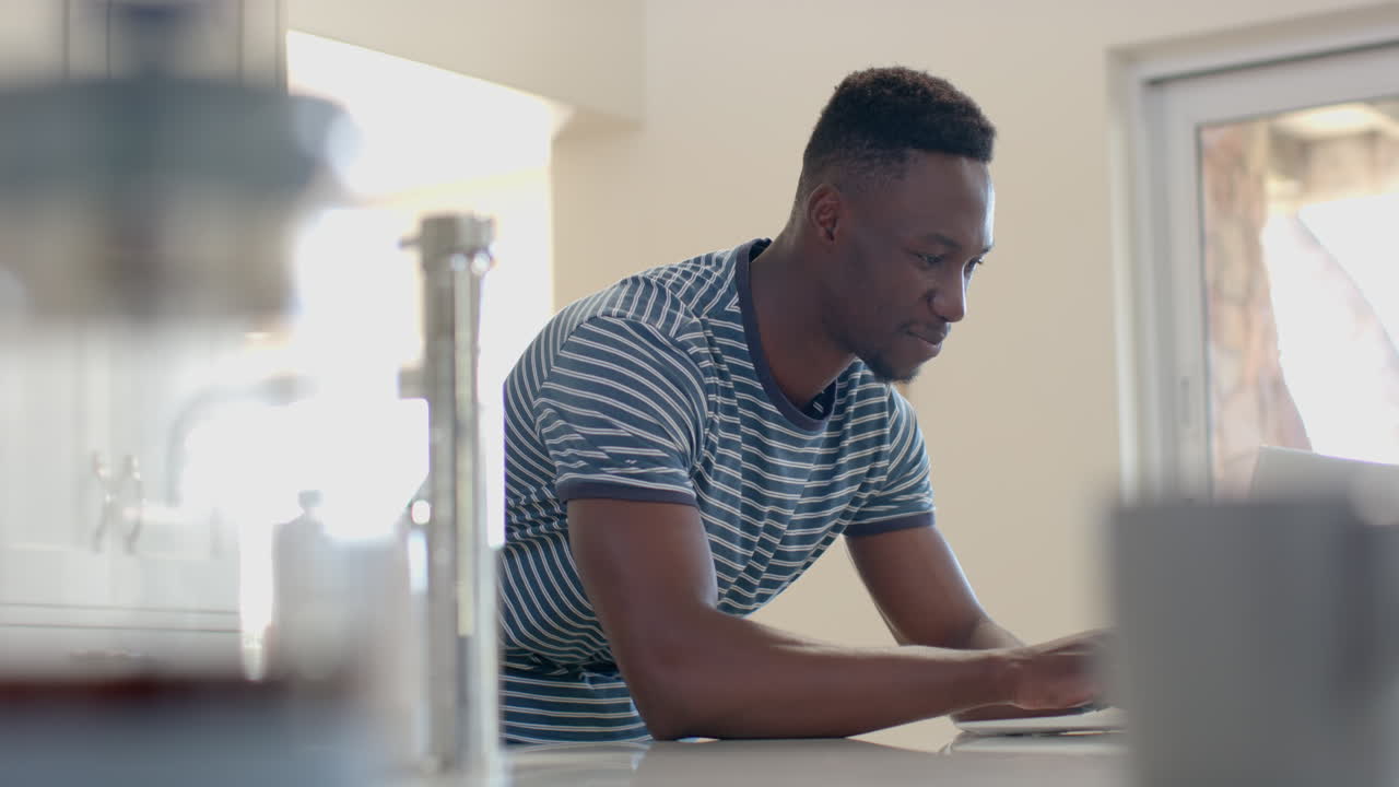 Happy african american man leaning on counter and using smartphone in sunny kitchen, slow motion