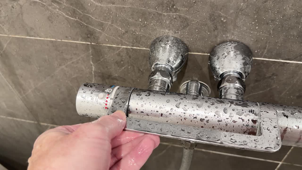 Nærbilde av en hånd som vrir på en termostatkran i krom på et vått dusjbatteri.