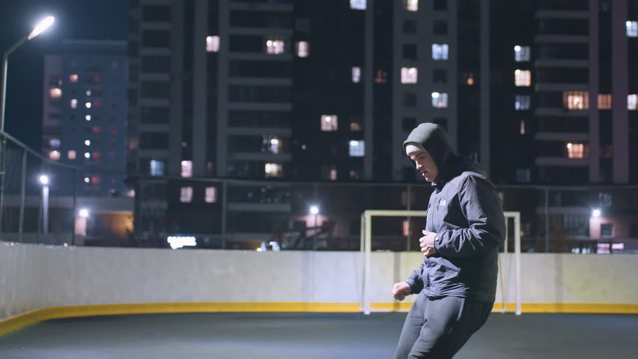 Side view of athlete in hoodie kicking soccer ball against wall under bright stadium lighting at night, urban cityscape with illuminated buildings and goalpost in background