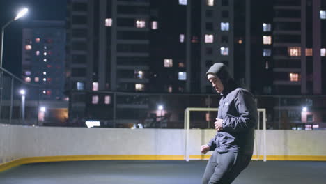 side view of athlete in hoodie kicking soccer ball against wall under bright stadium lighting at night, urban cityscape with illuminated buildings and goalpost in background
