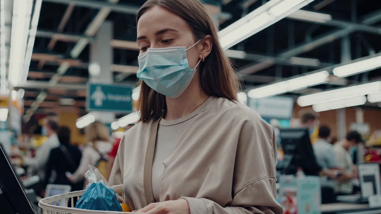 식료품 가게 환경에서 전염병 안전 조치 동안 새로운 쇼핑 규범을 나타내는 셀프 체크아웃 카운터에서 보호 얼굴 마스크를 입은 여성이 기다리고 있습니다.