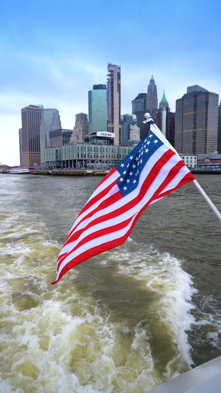 Bandiera americana che sventola da un traghetto con lo skyline di Manhattan, New York. Vista ravvicinata della bandiera sopra la scia con grattacieli e cielo coperto sullo sfondo.