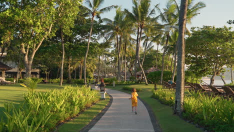 a serene stroll through vibrant green gardens lined with palm trees captures the tranquil essence of bali as young girl in yellow dress runs on path at sunrise