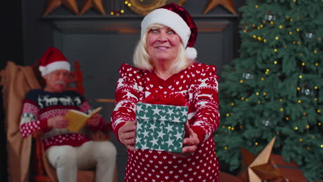 Mature-grandmother-in-New-Year-sweater-and-hat-presenting-Christmas-gift-box,-holidays-at-home