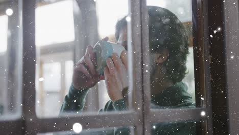 animation of window and snow falling over caucasian man drinking coffee at christmas