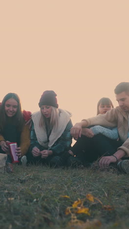 joyful young friend tourists sit at burning bonfire talking about dreams on meadow at sunset in autumn low angle shot