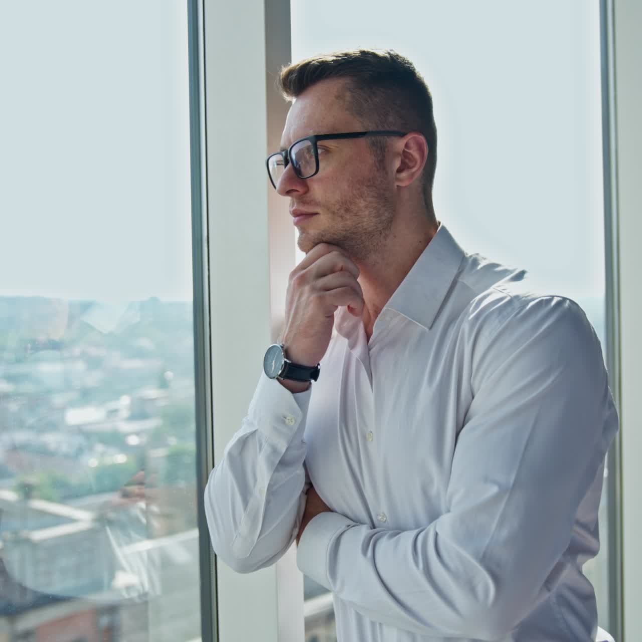 Thoughtful young adult man in glasses and white shirt stands leaning on the window frame. Entrepreneur thinking of something looking at the cityscape