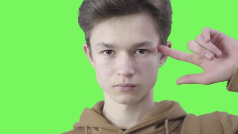 close-up of brunette caucasian boy twisting finger at temple. portrait of serious teenager showing crazy nuts gesture. misunderstanding, astonishment, misinterpretation. chromakey, green screen.