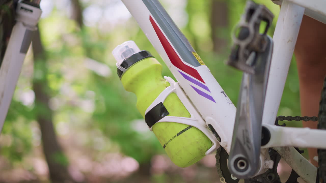 Closeup Of Bicycle Drinking Bottle, Stationary Bicycle With Bright Container Amid Muddy Trail Scene, Detailed View Of Bicycle With Vibrant Water Bottle Secured Within White Cage On Frame