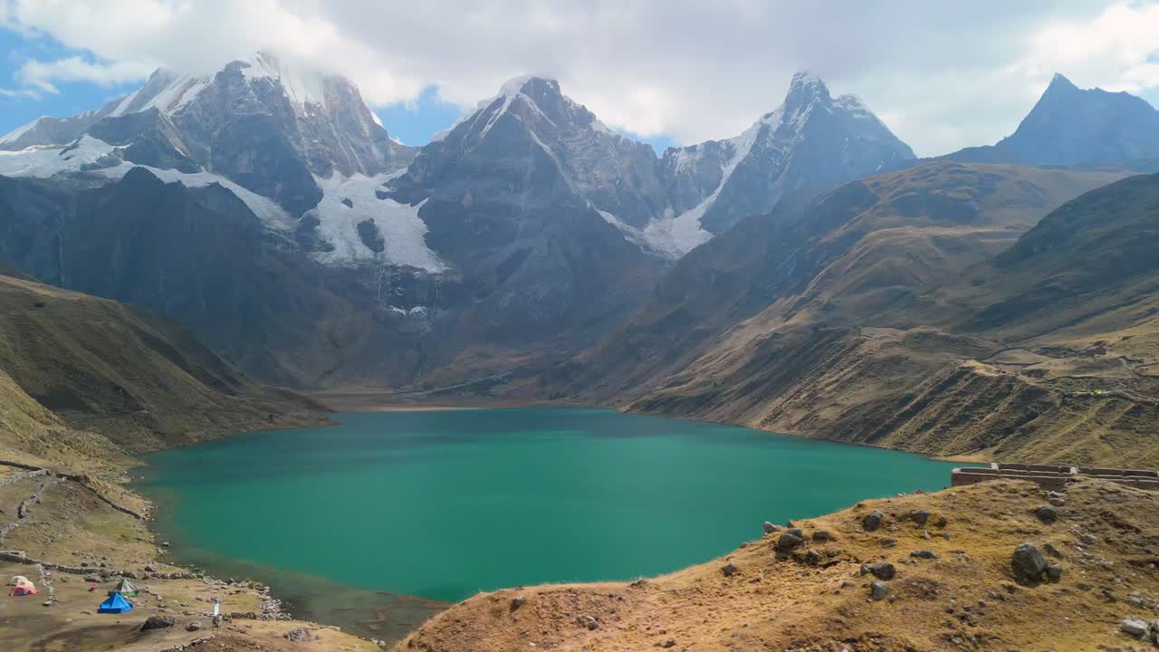 A stunning aerial shot reveals the turquoise Laguna Jahuacocha and a remote campsite, with the majestic Yerupajá and Jirishanca peaks of the Huayhuash range in the background