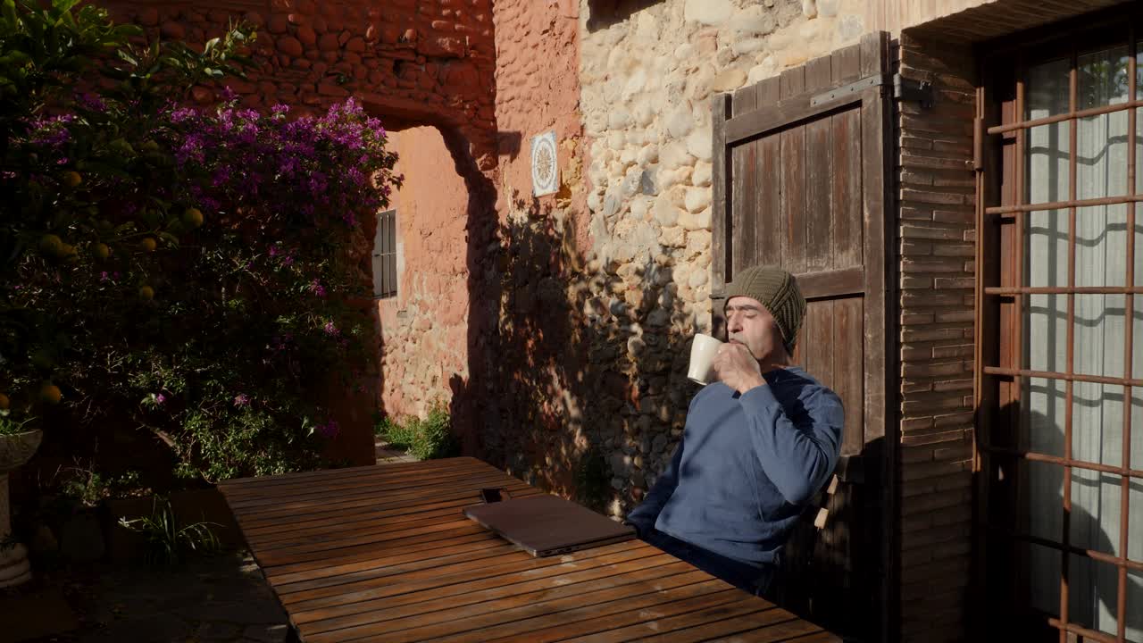 un hombre cierra su computadora portátil afuera de una cabaña y bebe café disfrutando del sol