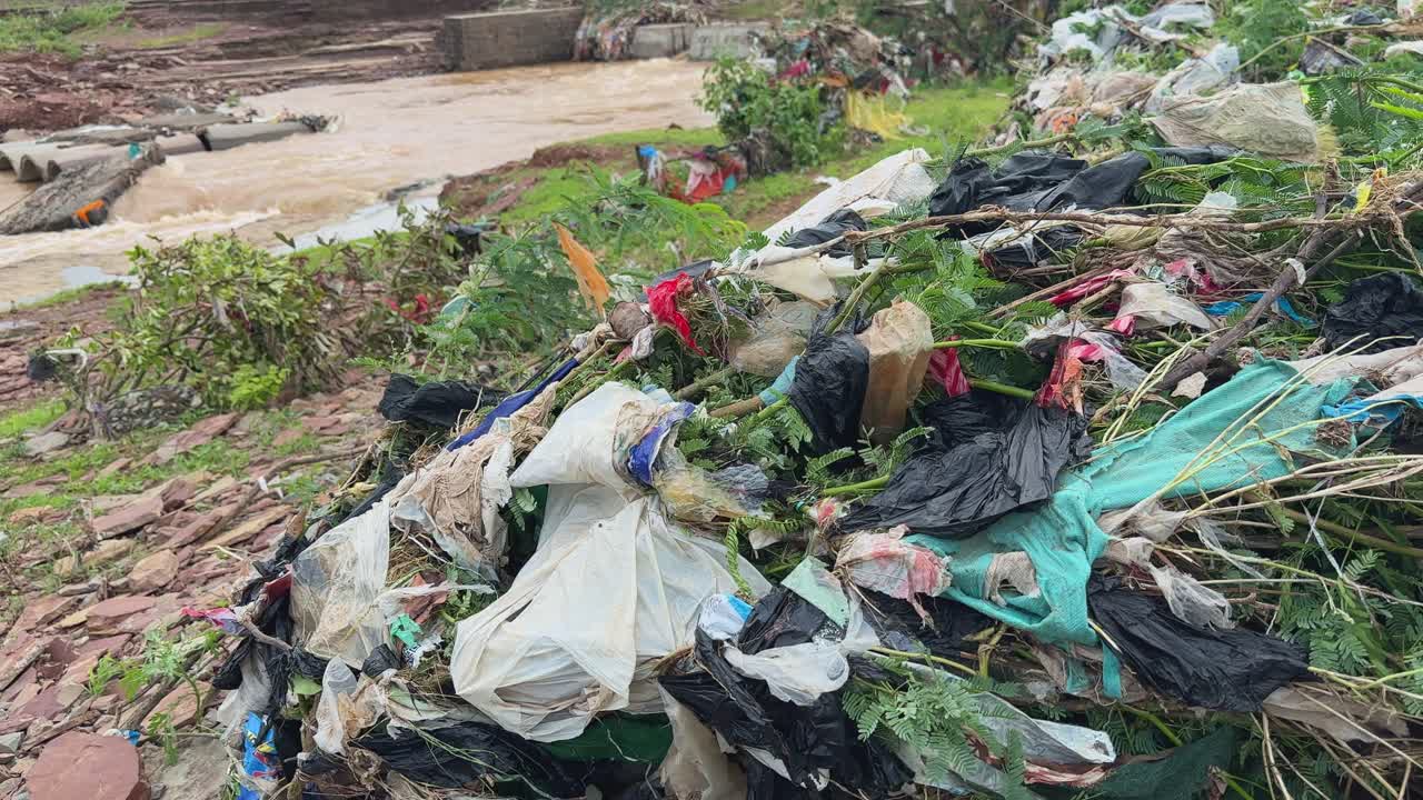 Push in shot of a large accumulation of trash and debris, primarily plastic, along the banks of a flowing river after the flooding happened
