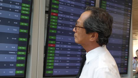 A-Japanese-businessman-stands-at-Ben-Gurion-Airport-in-Israel,-looking-at-the-arrivals-and-departures-board-He-is-photographed-from-his-left-side-with-the-arrivals-and-departures-board-in-front-of-him