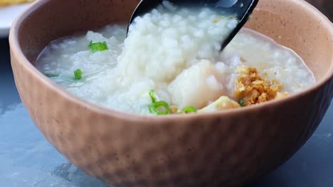a black ladle stirs and serves porridge garnished with green onions and crispy toppings in a textured brown bowl.