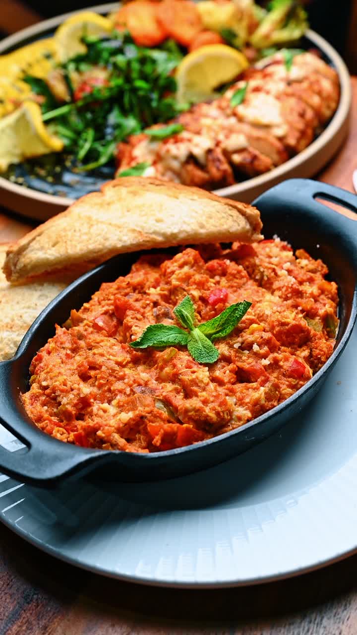 Apparecchiatura della tavola con uova menemen e sfondo di un piatto di pollo alla griglia. Due piatti diversi che includono uova strapazzate in padella e pollo alla griglia con verdure su un tavolo di legno di un ristorante.