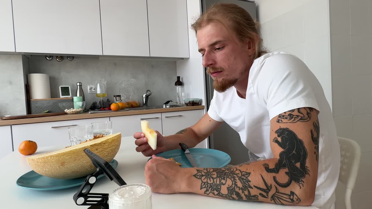Junger Mann mit Tattoos isst frische Melone in einer modernen Küche.