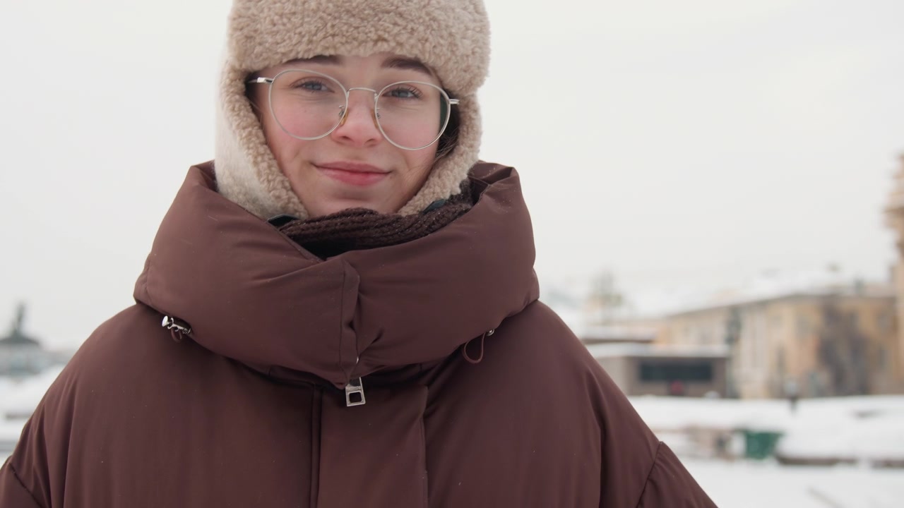 Primo piano medio mostra una persona che indossa un cappotto trapuntato marrone oversize con collo alto e un cappello peloso beige, rivolta verso la fotocamera in una luce nevosa e nuvolosa, occhiali trasparenti visibili e un sorriso sottile che cambia da