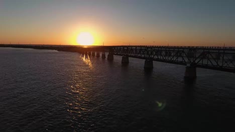 AERIAL:-Pulling-away-from-an-old-broken-down-bridge-at-sunset