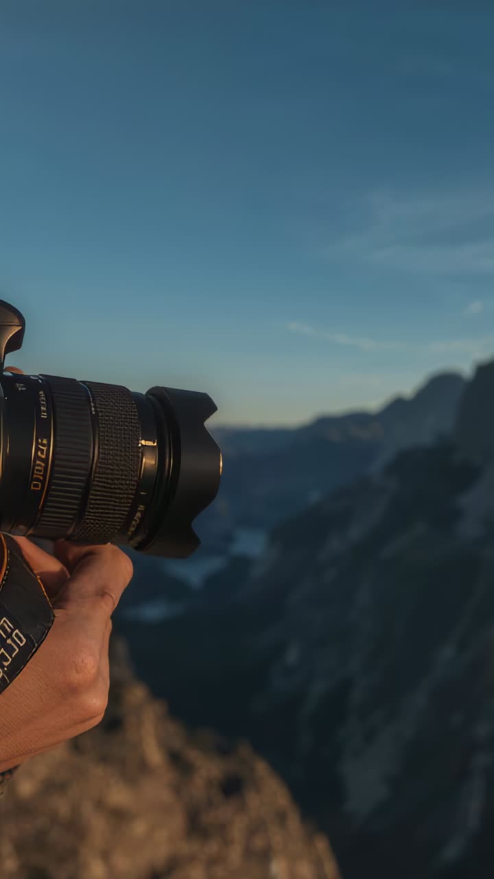 Pystyvideo: Kuvakoko, jossa ammattilainen pitää oranssilla hihnalla varustettua DSLR-kameraa reunan päällä aamunkoitteessa, laaksossa ja huipuilla, tilaa tekstille