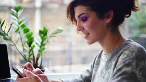 Vista-De-Cerca-De-Una-Hermosa-Joven-Usando-Su-Teléfono-Móvil-En-La-Cafetería-Y-Sonriendo.-Mujer-Usando-La-Aplicación-En-El-Teléfono-Inteligente-En-La-Cafetería-Bebiendo