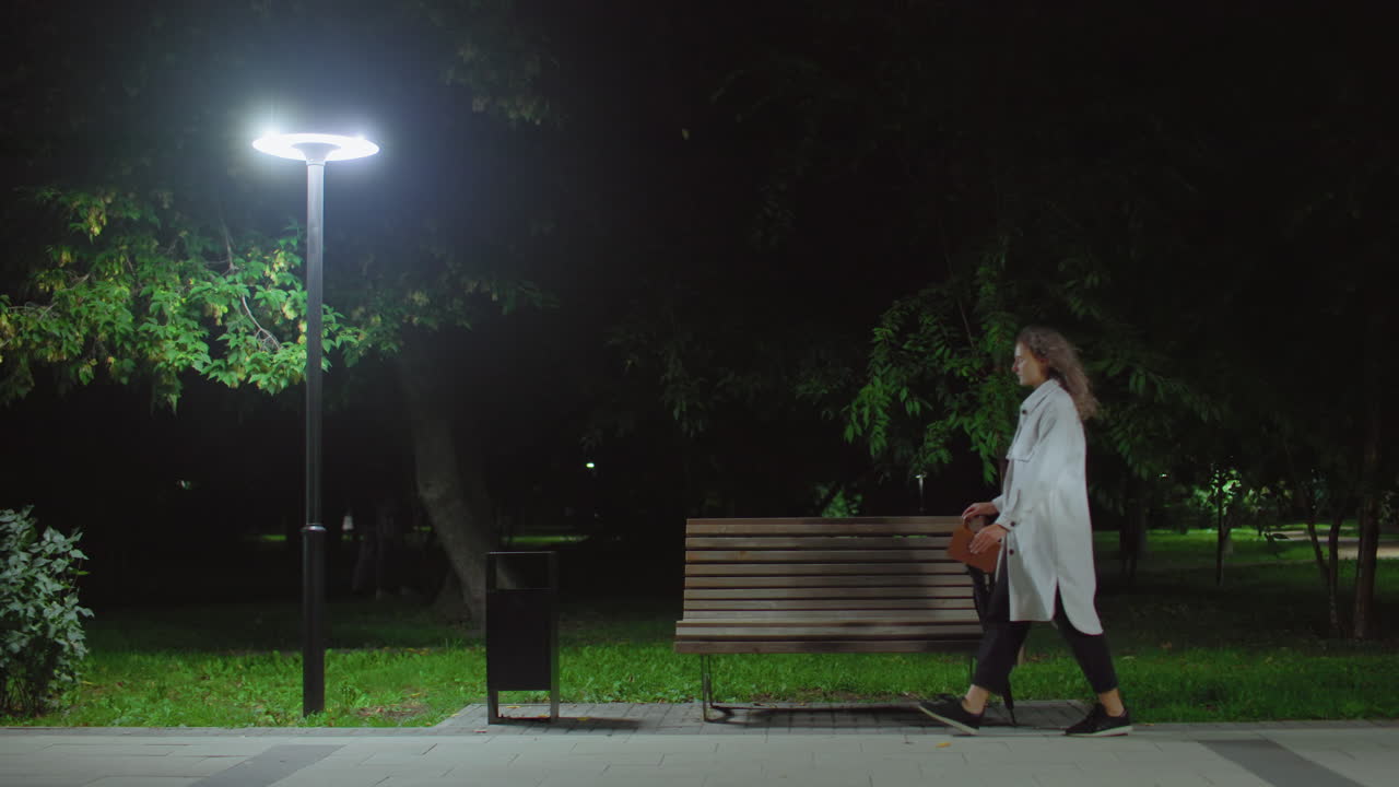 Young lady walks to bench under glowing streetlight sits down drops umbrella opens book to read while people move in dark background creating peaceful calm night atmosphere in green park