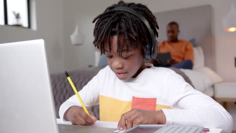 Niño-Afroamericano-Usando-Una-Computadora-Portátil-Para-Una-Lección-En-Línea-Con-Su-Padre-En-Segundo-Plano-En-Casa,-Cámara-Lenta