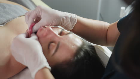 lower lips of young lady gently pressed and smoothed by wellness technician wearing protective gloves during facial massage session under bright lamp conveying calm care and precision touch