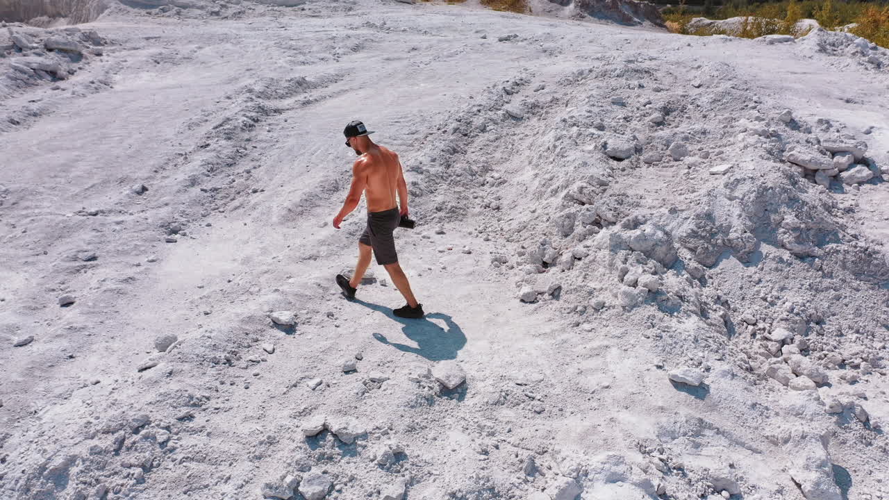 Strong man in white canyon. Healthy man without shirt walking on a rocky background in summer. Aerial view on a sporty male in cap and shorts in nature.