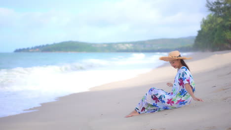 a young woman holds on to her sun hat as she sits on the beach in her coverup