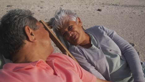 Happy-senior-hispanic-couple-talking-and-relaxing-on-sunbeds-on-beach-at-sunset