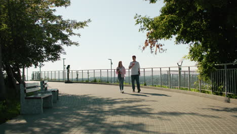 siblings walking near iron railing, returning from class and engaging in happy conversation, while another person walks in the distance under bright sunlight with benches, in the background