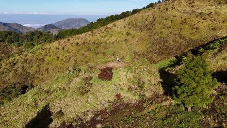 drone footage capturing a lone figure on green mountaintop surrounded by ridges, dense forests, and towering mountain in the distance. breathtaking moment of solitude and nature