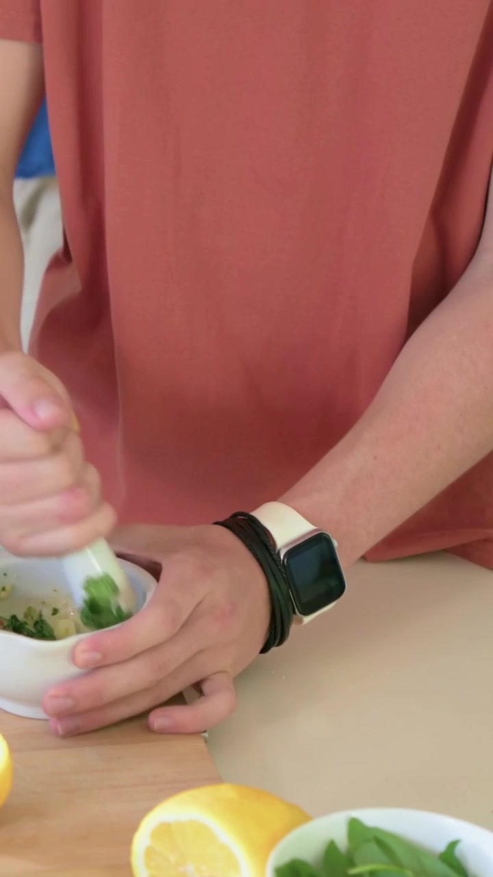 Vertical video: Adding lemon man coral tee mashing herbs mortar in kitchen grabbing oil, copy space