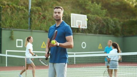 hombre guapo entrenando y golpeando la pelota con raqueta mientras su familia juega al tenis en el fondo