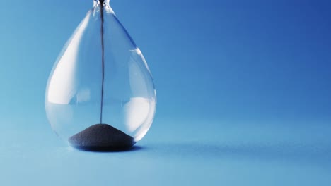 Video-of-close-up-of-hourglass-with-black-sand-pouring-with-copy-space-on-blue-background