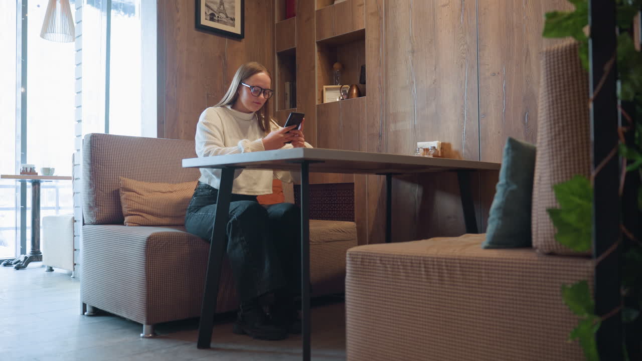 freelancer wearing glasses and white sweater seated in modern cafe booth chatting over phone with relaxed posture, surrounded by wooden walls, indoor plants, and daylight from window