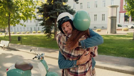 Happy-guy-with-long-curly-hair-in-a-denim-shirt-and-a-white-motorcycle-helmet-hugs-his-brunette-girlfriend-in-a-checkered-shirt-Near-his-motorcycle-in-a-summer-city-park