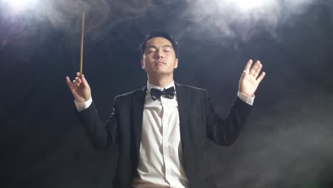 asian conductor man holding a baton closing his eyes and showing gesture in the black studio with fog