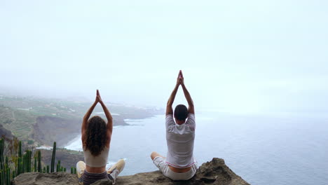 Positioned-atop-a-mountain,-a-man-and-woman-meditate-on-rocks,-raising-hands-in-unison,-absorbing-the-ocean-vista,-and-performing-relaxing-breaths